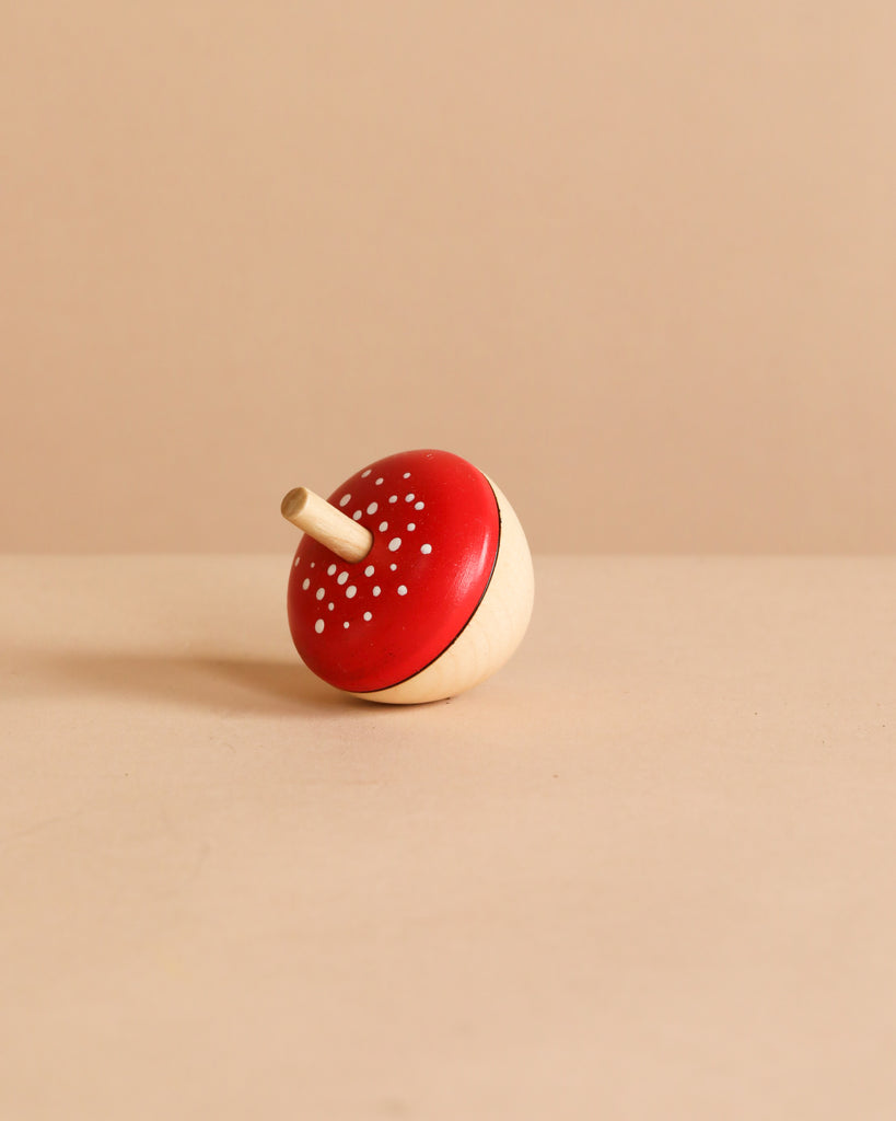 An Egg-Shaped Spinning Top with a red, mushroom-like cap and white dots sits on a beige surface against a plain background, crafted to meet toy safety standards.