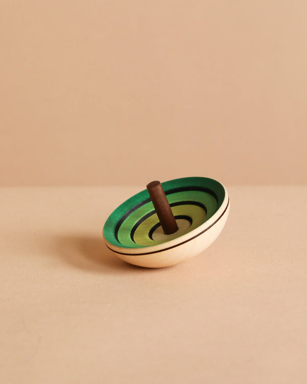 The Mader UFO Flora Spinning Top - Green, crafted from maple wood with green concentric circles and a brown handle, is displayed on a beige surface against a light beige background.