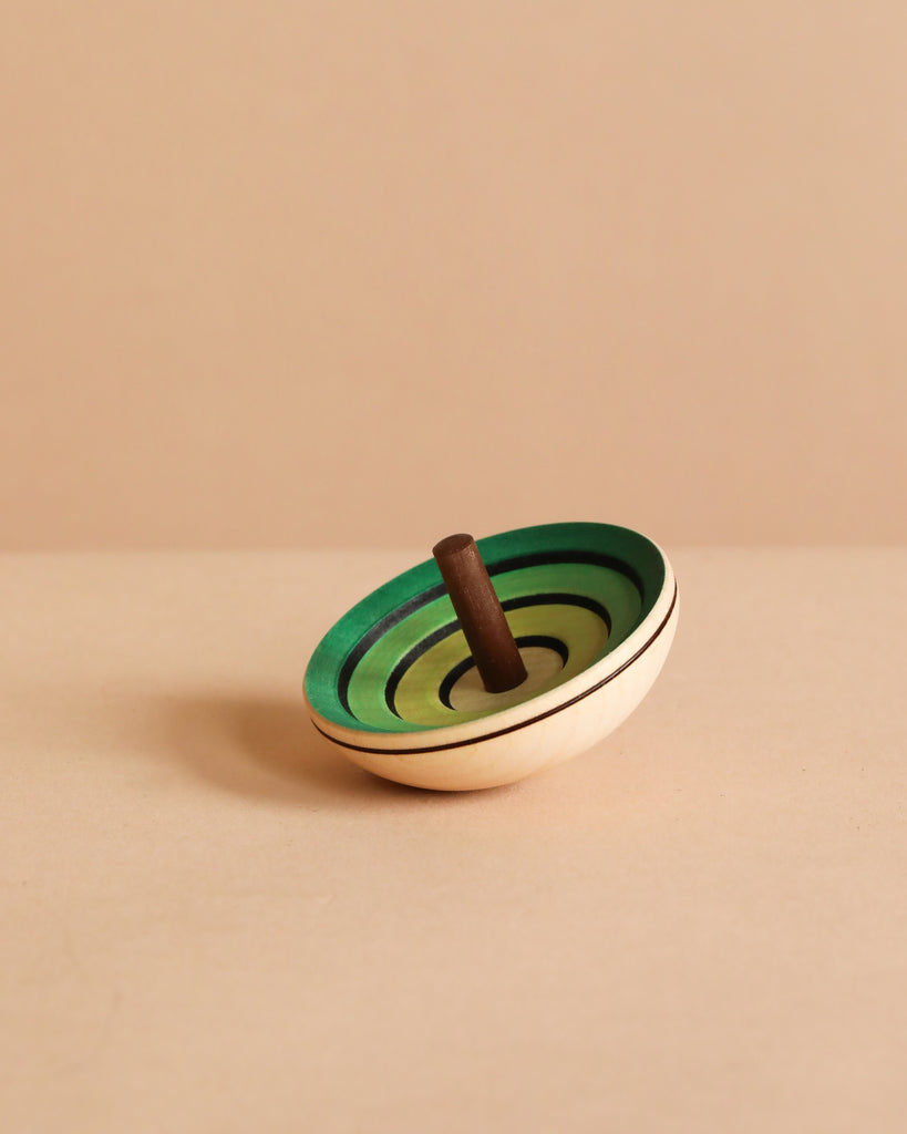 The Mader UFO Flora Spinning Top - Green, crafted from maple wood with green concentric circles and a brown handle, is displayed on a beige surface against a light beige background.
