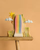 The Bumbu Handmade Wooden Rainbow Cliff is a colorful, Waldorf-inspired sculpture featuring a radiant sun, white clouds, and painted trees on a wooden base set against a beige background.