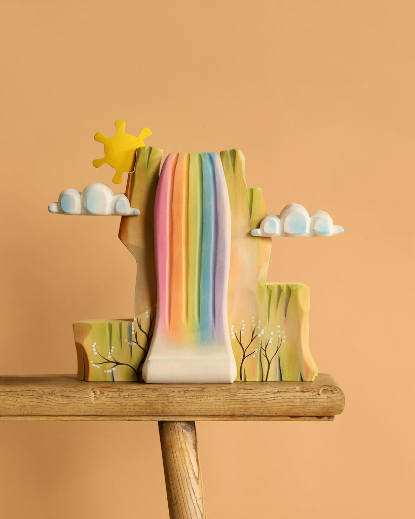 The Bumbu Handmade Wooden Rainbow Cliff is a colorful, Waldorf-inspired sculpture featuring a radiant sun, white clouds, and painted trees on a wooden base set against a beige background.
