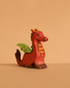 The Bumbu Handmade Wooden Baby Dragon features a small, red body with green wings and orange accents, displayed on a beige surface that complements its fantasy-inspired design—ideal for imaginative play.