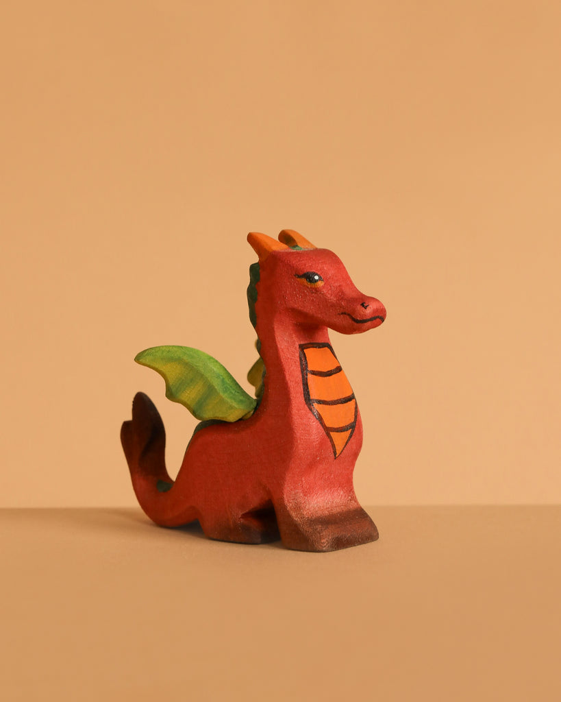 The Bumbu Handmade Wooden Baby Dragon features a small, red body with green wings and orange accents, displayed on a beige surface that complements its fantasy-inspired design—ideal for imaginative play.