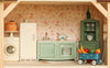 A miniature kitchen from the Maileg Fully Furnished Farmhouse Set, featuring a white washing machine topped with a layer cake, a refrigerator, a green stove unit with a sink, a green hutch with glassware, and a blue toy wagon with vegetables.