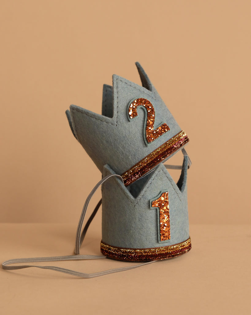 Two small Storm Gray Felt Crowns with elastic bands are stacked together, ideal for birthdays. Each crown features a glittery "1" on the front and a "2" on the back, outlined in gold and copper on a beige background.