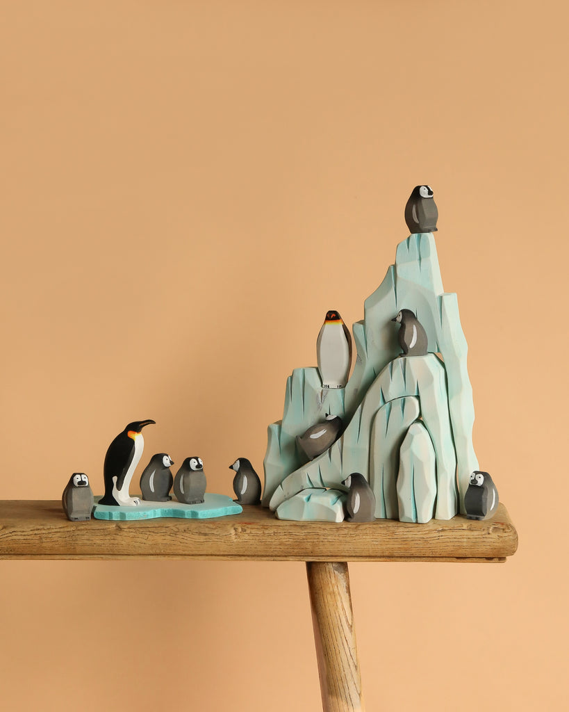 The Bumbu Handmade Wooden Penguin Set features small penguin figurines and an adult arranged by a light blue iceberg on a wooden surface, ideal for Waldorf-inspired play or creating a handmade Antarctica scene.