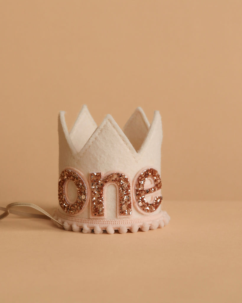 The Linen and Rose Crown is a small white felt crown featuring glittery gold "one" letters, perfect for birthday celebrations. It adds rose gold flair to your little one's big day and stands out on a neutral beige background.