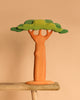 The Bumbu Handmade Wooden Extra Large Baobab Tree, featuring a thick brown trunk and layered green foliage, sits on a wooden surface against a beige background.