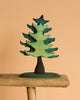 The Bumbu Handmade Wooden Extra Large Green Spruce Tree, featuring a stylized design with green and blue hues and a dark trunk, is displayed on a rustic wooden plank against a plain beige background, highlighting its handcrafted charm.
