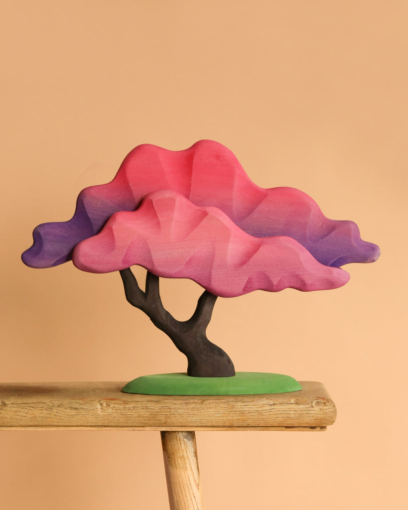 The Bumbu Handmade Wooden Japanese Maple Tree, with pink and purple layered foliage, is displayed on a wooden stool against a plain beige background.