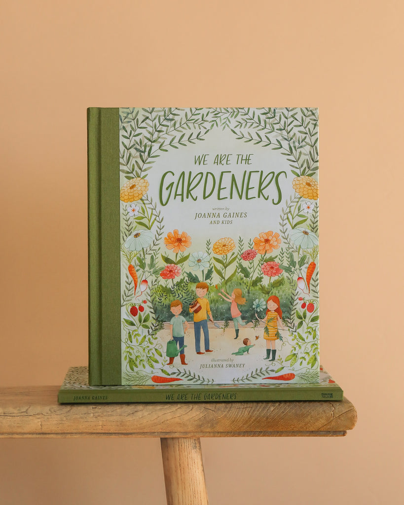 We Are The Gardeners," a children's book, stands upright on a wooden bench with another copy below it against a beige background. The cover features illustrated kids learning from their family's thriving garden.