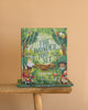 The children's book "The World Needs The Wonder You See" is displayed upright on a wooden stool against a peach background, inviting kids to explore an imaginative adventure filled with illustrated children, animals, and plants.