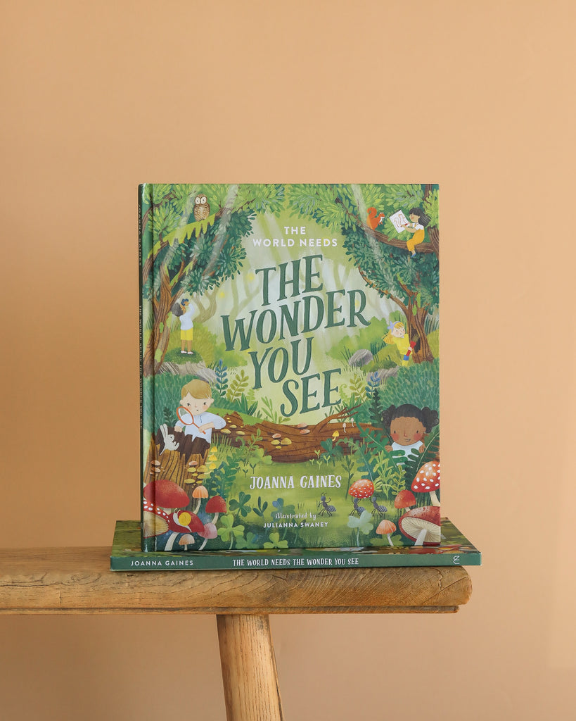 The children's book "The World Needs The Wonder You See" is displayed upright on a wooden stool against a peach background, inviting kids to explore an imaginative adventure filled with illustrated children, animals, and plants.