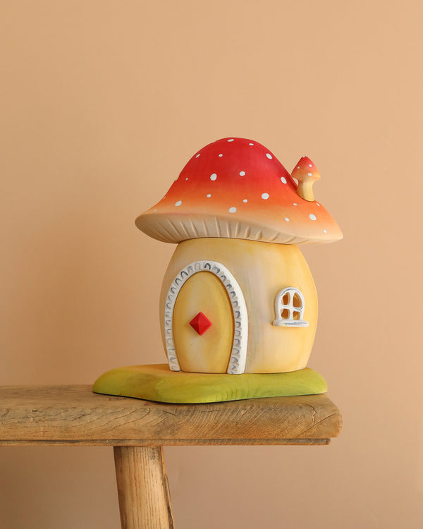 The Bumbu Wooden Toadstool House, designed as a red and white spotted mushroom with an arched door and window, rests on a rustic bench made from sustainably sourced wood against a plain beige background.