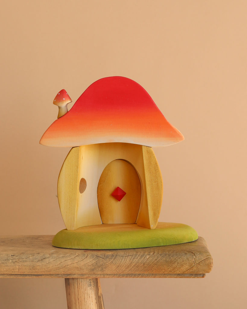 The Bumbu Wooden Toadstool House is a small, handcrafted playset piece with a red and orange roof, tiny chimney, and green base, all made from sustainably sourced wood. It rests on a rustic surface with a plain beige background.