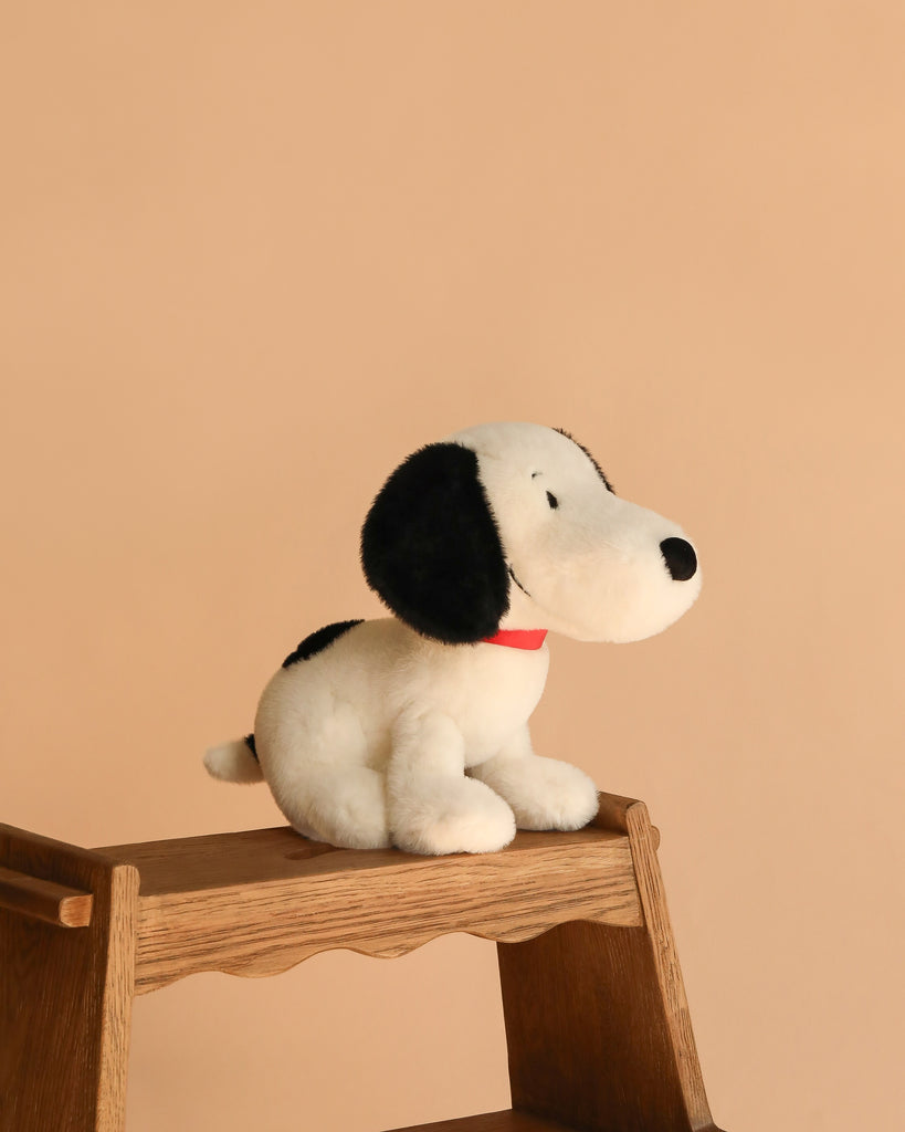 A Plush Peanuts Snoopy Good Ol' Charlie Brown with black ears, a black spot, and a red collar sits on a wooden step stool against a plain beige background.