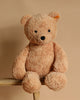 The Steiff XL Jimmy Teddy Bear, standing 22 inches tall and flaunting light brown fur, rests on a wooden stool against a beige backdrop. Its black eyes, signature Button in Ear, and small tag enhance its timeless charm.