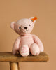 The 10-inch My First Steiff Pink Teddy Bear, featuring a yellow tag and "My first Steiff" pink embroidery on its left foot, sits on a wooden stool against a beige background—an adorable baby gift.