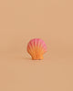 A pink and orange seashell-shaped object is centered against a plain, light beige background, echoing the style of the Bumbu Handmade Wooden Ocean Set.