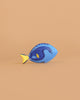 A small fish figurine from the Bumbu Handmade Wooden Ocean Set, featuring a blue body and yellow tail with a simple, cartoon-like design, is displayed on a light brown background.
