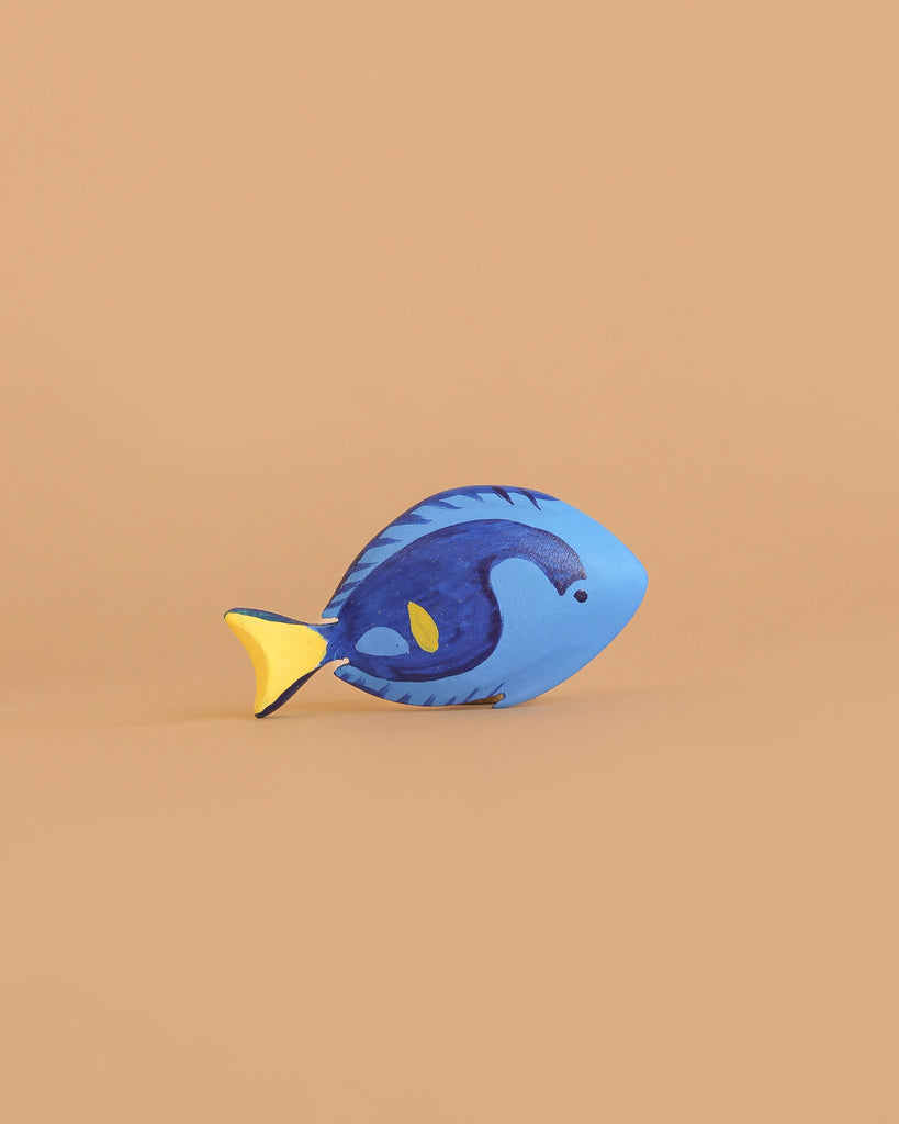 A small fish figurine from the Bumbu Handmade Wooden Ocean Set, featuring a blue body and yellow tail with a simple, cartoon-like design, is displayed on a light brown background.