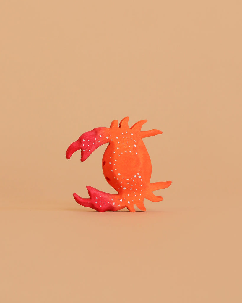A handmade orange and pink crab with white dots from the Bumbu Wooden Ocean Set is centered on a plain beige background.