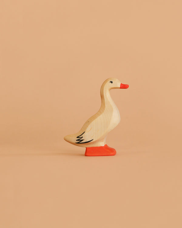 The Bumbu Handmade Wooden Domestic Duck is a small, handcrafted toy featuring a beige body, orange beak, and orange feet, displayed on a solid light peach background.
