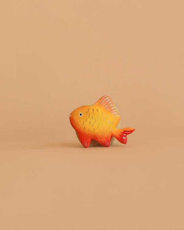 The Bumbu Handmade Wooden Golden Fish, featuring orange and yellow hues with red fins, is displayed against a simple, light brown background.