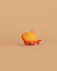 A small, orange and yellow toy fish with red fins sits on a light brown background—a charming piece from the Bumbu Handmade Wooden Ocean Set.