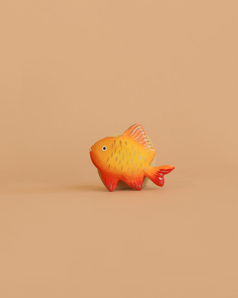 A small, orange and yellow toy fish with red fins sits on a light brown background—a charming piece from the Bumbu Handmade Wooden Ocean Set.
