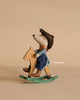 A Maileg Cowboy Set features a mouse in a blue checkered shirt and hat riding a wooden rocking horse, set against a beige background.