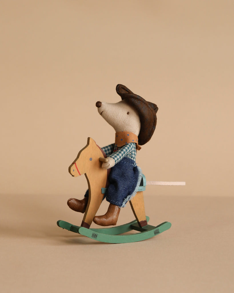 A Maileg Cowboy Set features a mouse in a blue checkered shirt and hat riding a wooden rocking horse, set against a beige background.