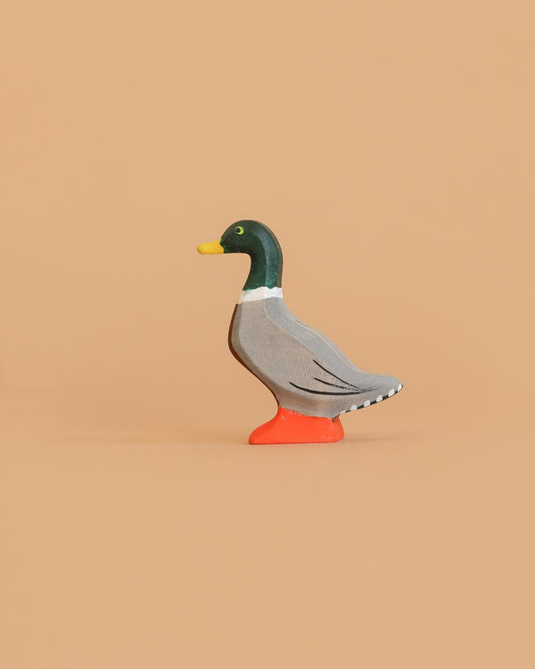 The Bumbu Handmade Wooden Mallard Duck, featuring a green head, yellow beak, gray body, and orange feet, stands on a plain beige background. This charming figurine is crafted from sustainable maple wood.