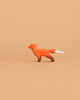 The Bumbu Handmade Wooden Fox Cub Curious, a small orange minimalist toy with a white-tipped tail, stands on a plain light brown background.