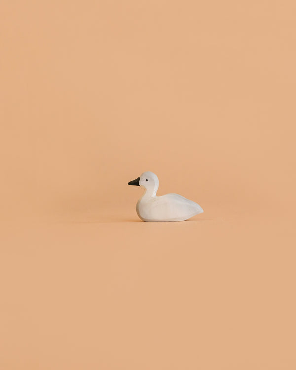 The Bumbu Handmade Wooden Baby Swan, a small white handcrafted wooden toy with a black beak, sits on a plain, light peach background.