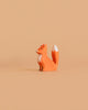 The Bumbu Handmade Wooden Fox Cub Sitting shows a small, orange, handcrafted wooden fox with a white-tipped tail and chest, displayed on a plain light brown background.