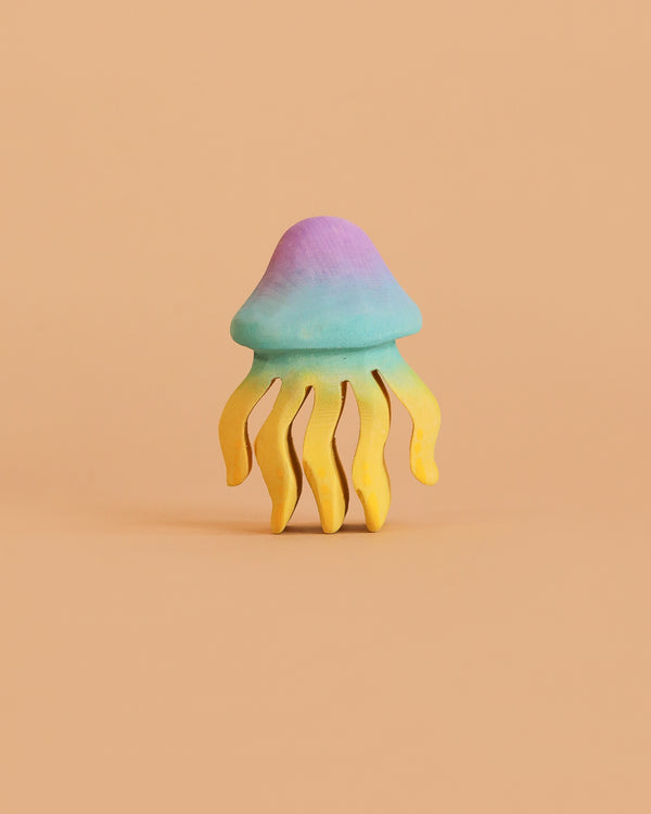The Bumbu Handmade Wooden Jellyfish, a small pastel sculpture with a blue-purple dome and yellow wavy tentacles, creates an enchanting eco-friendly toy when displayed against a plain peach background.