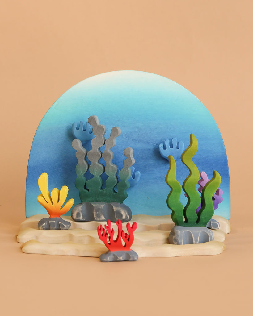 A colorful wooden diorama features blue, green, yellow, and red seaweed and coral on a blue and tan background—perfect to pair with the Bumbu Handmade Wooden Ocean Set.