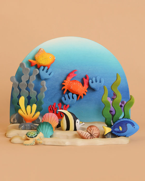 The Bumbu Handmade Wooden Ocean Set, featuring crab and turtle figures, is displayed in an underwater scene with a blue background, sandy foreground, and peach backdrop alongside colorful clay sea creatures.
