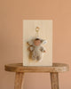 The Olli Ella Cozy Bag Charm - Lamby Pip, featuring sheep ears and a gray outfit, hangs on a wooden panel displayed atop a rustic stool against a plain beige background.