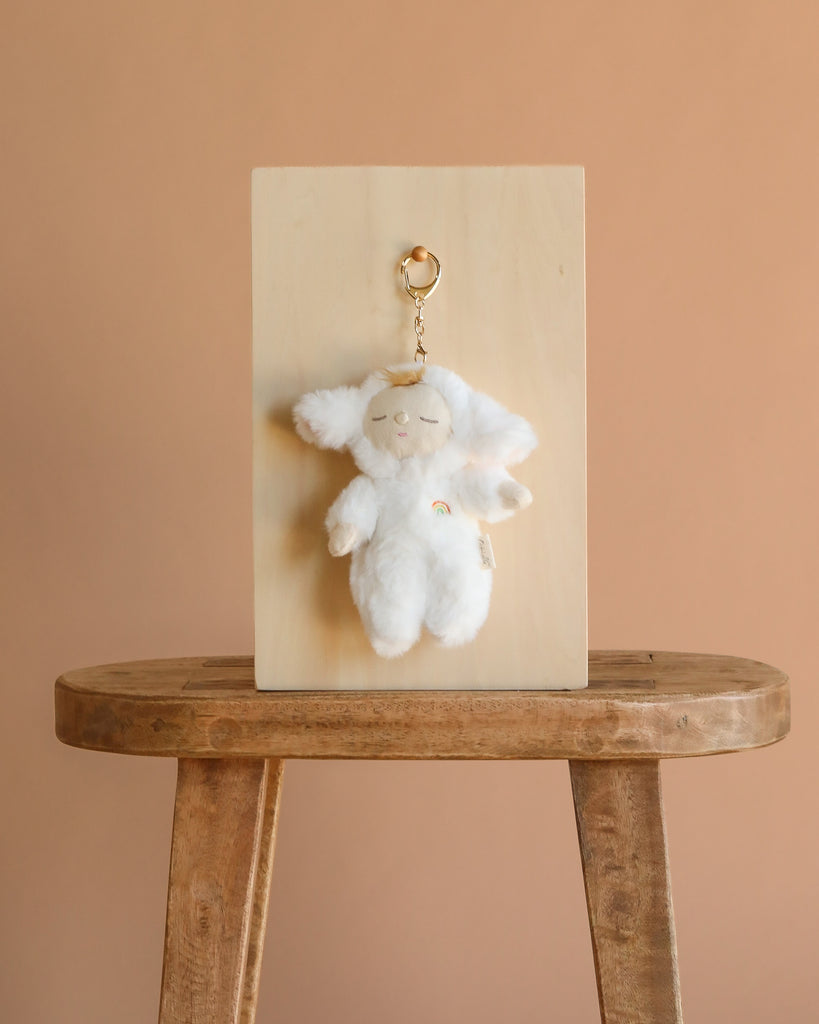 The Olli Ella Cozy Bag Charm - Lamby Pookie, a fluffy white plush keychain with floppy ears and a rainbow patch, hangs on a wooden board set on a stool against a light brown background.