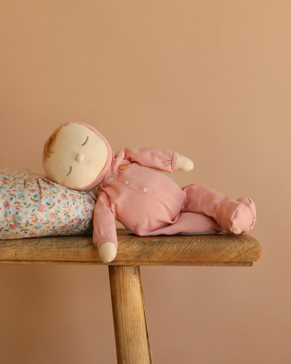 The Olli Ella Dozy Dinkum - Moppet, a soft cotton baby doll in a pink outfit, rests on its side on a wooden bench with its head on a floral pillow against a peach-colored background.