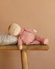 The Olli Ella Dozy Dinkum - Moppet, a soft cotton baby doll in a pink outfit, rests on its side on a wooden bench with its head on a floral pillow against a peach-colored background.
