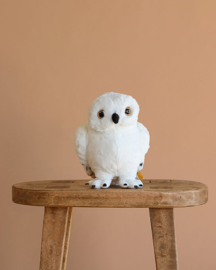 The Steiff Harry Potter "Hedwig" Owl 8" plush, featuring yellow eyes, sits on a wooden stool against a beige background—an ideal gift for any Harry Potter fan.