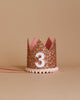 The Rose Gold Glitter Crown rests elegantly against a beige background, adorned with shimmering gold glitter and showcasing a white number "3" on the front. The crown's base is trimmed with delicate white pom-poms, while pink ribbon ties extend from the sides, making it perfect for birthday celebrations or party hats.