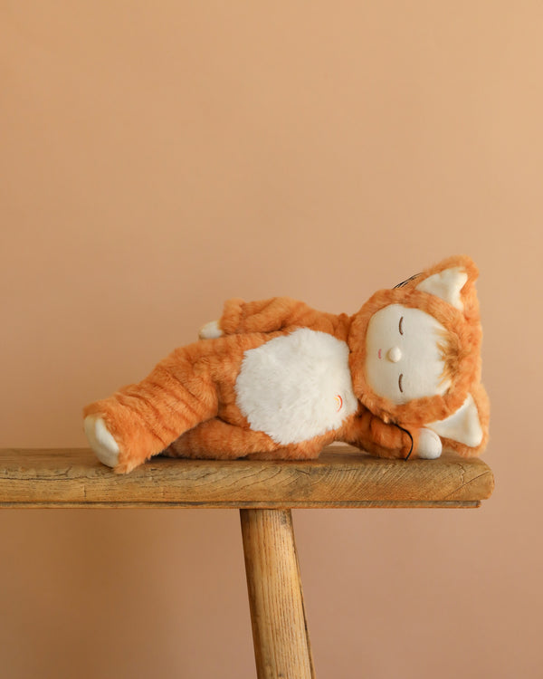 The Olli Ella Cozy Dinkum Doll – Tabby Cat Jinx is shown lying on its side, as if asleep, on a wooden bench with a plain beige background.