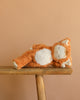 The Olli Ella Cozy Dinkum Doll – Tabby Cat Jinx is shown lying on its side, as if asleep, on a wooden bench with a plain beige background.