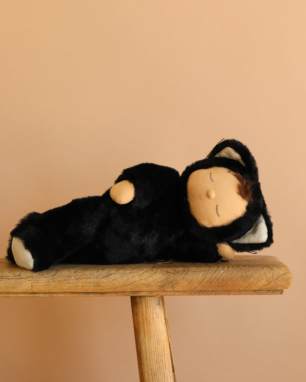 The Olli Ella Cozy Dinkum Doll - Black Cat Nox, a collectible plush in a black cat costume, rests on its side with closed eyes on a wooden bench against a plain beige background.