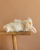 The Olli Ella Cozy Dinkum - Bunny Moppet is a collectible plush toy lying on its side in a soft white bunny costume with floppy ears, displayed on a wooden bench against a plain beige background.