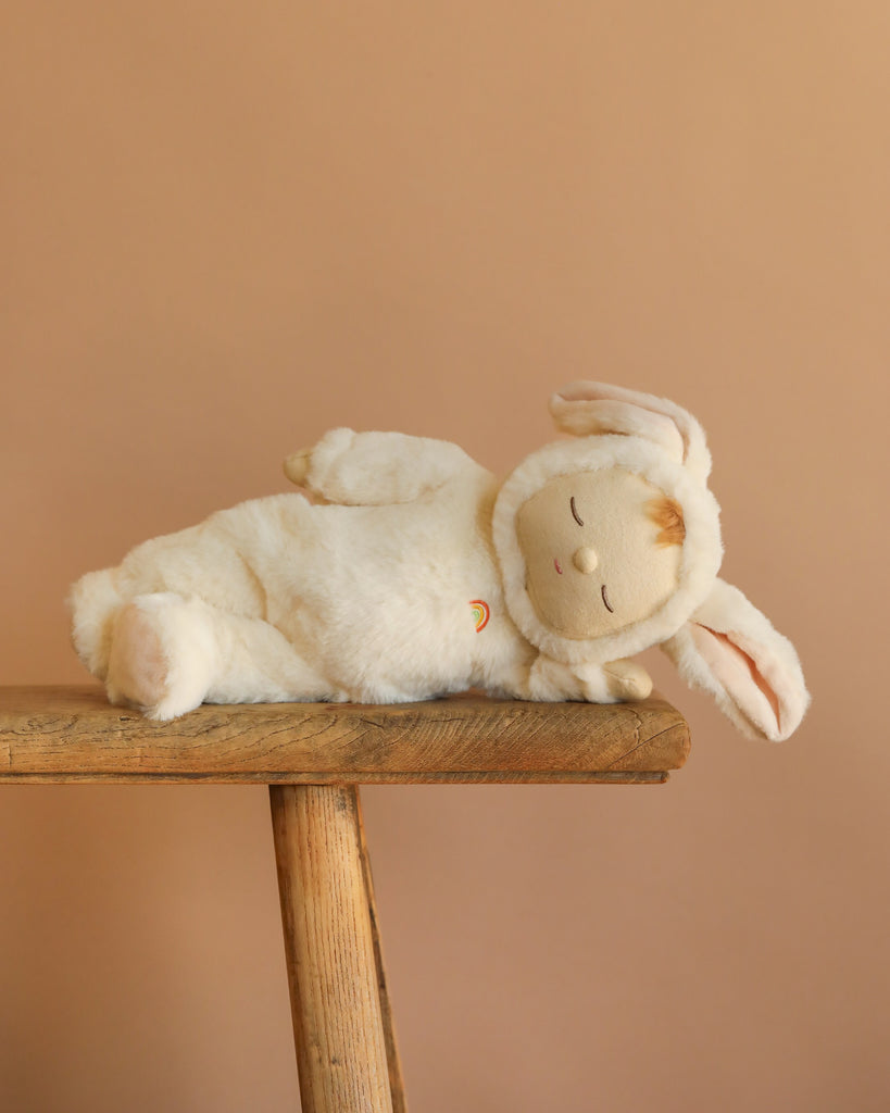 The Olli Ella Cozy Dinkum - Bunny Moppet is a collectible plush toy lying on its side in a soft white bunny costume with floppy ears, displayed on a wooden bench against a plain beige background.